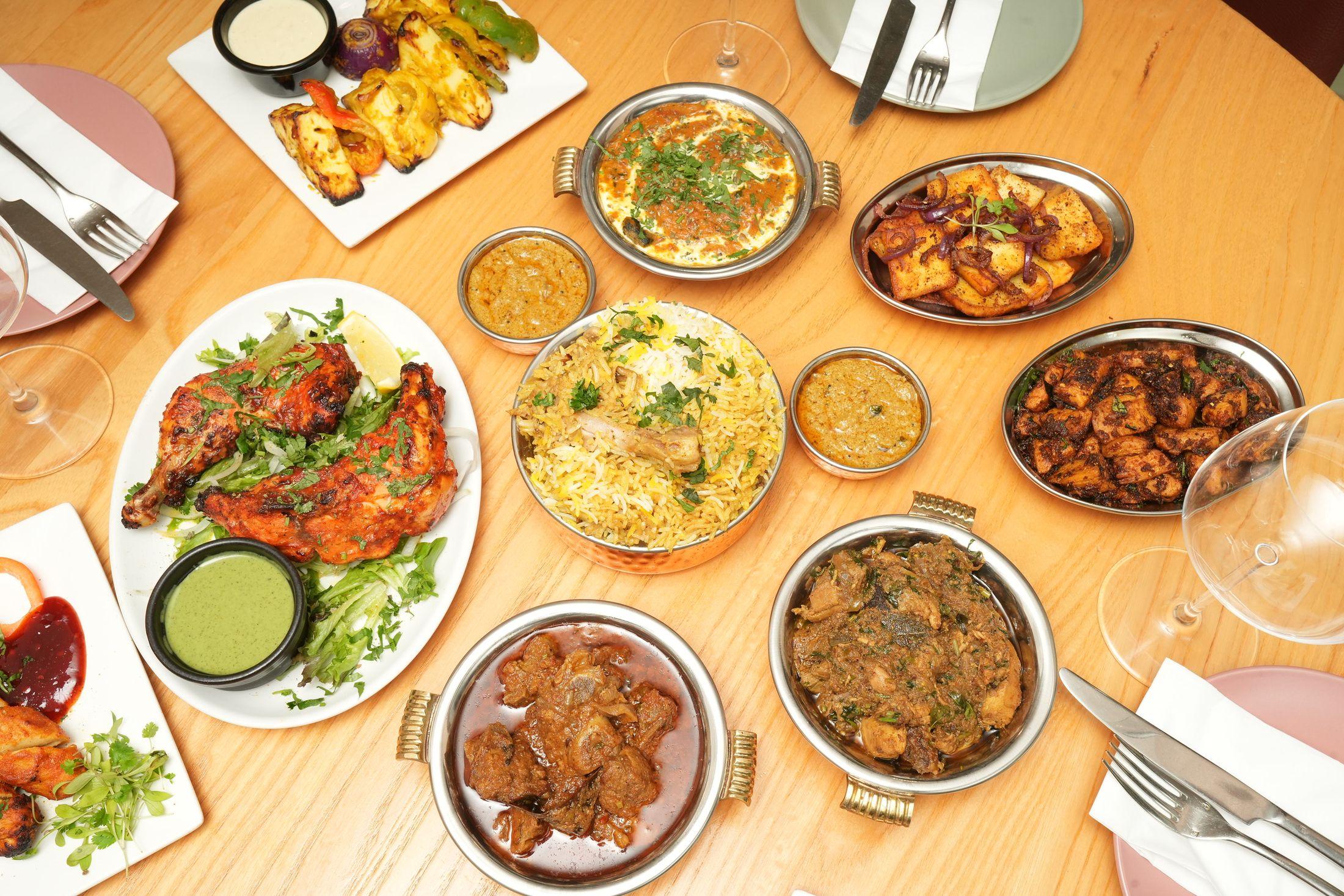 Overhead spread of Aara dishes — biryani in a large pot, tandoori chicken, curries and breads on a wooden table